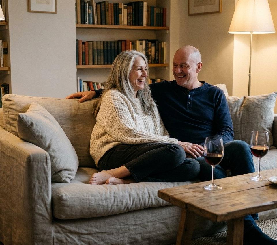 A couple in their fifties relaxing and laughing together on a sofa in a softly lit living room.